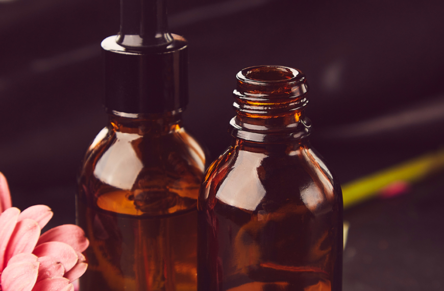 Botellas de vidrio ámbar con sérum facial y una pipeta sobre fondo oscuro, acompañadas de una flor rosada, representando productos de cuidado facial natural.