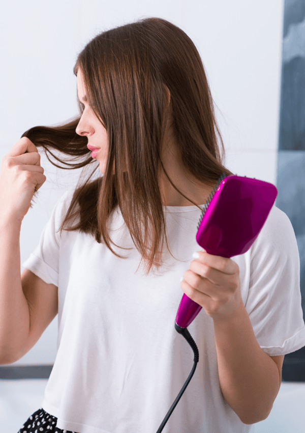 Mujer utilizando cepillos alisadores eléctricos de color rosa mientras examina un mechón de su cabello.