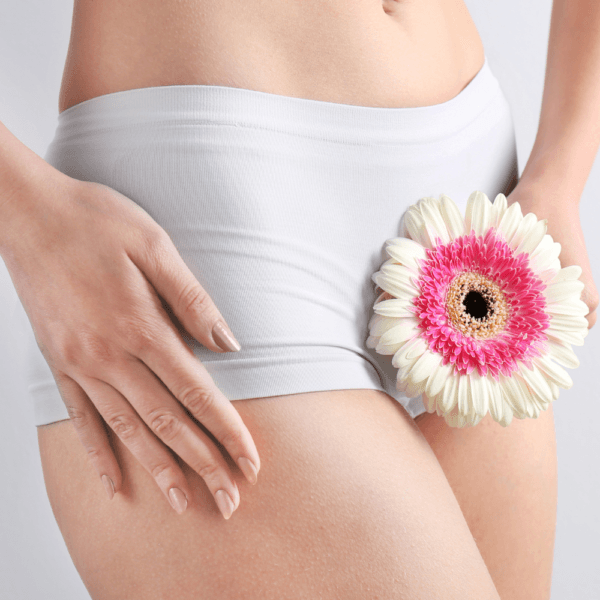 Mujer con ropa interior blanca sosteniendo una flor rosa y blanca sobre su cadera, con fondo claro.