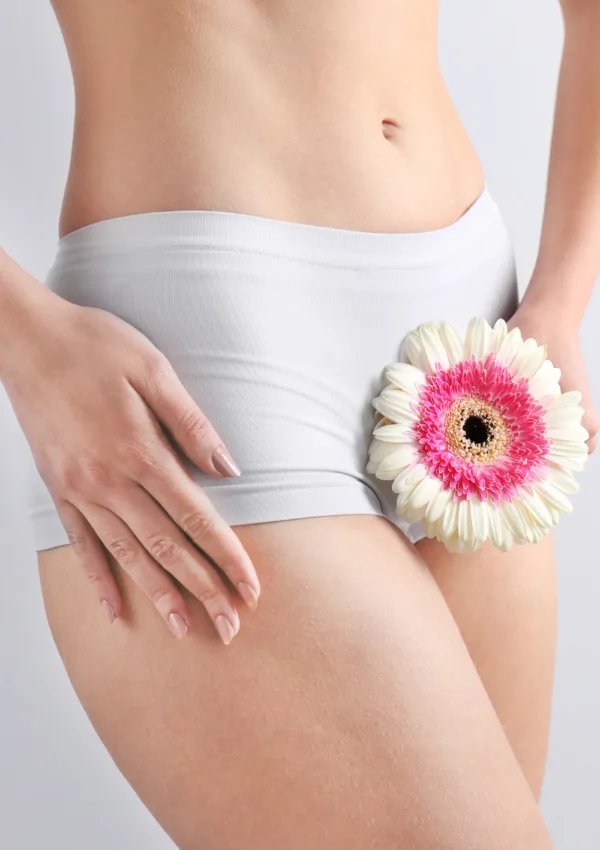 Mujer con ropa interior blanca sosteniendo una flor rosa y blanca sobre su cadera, con fondo claro.