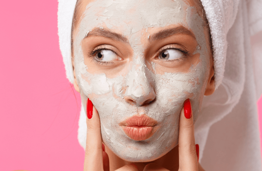 Mujer con mascarilla facial de arcilla y toalla en la cabeza sobre fondo rosa.